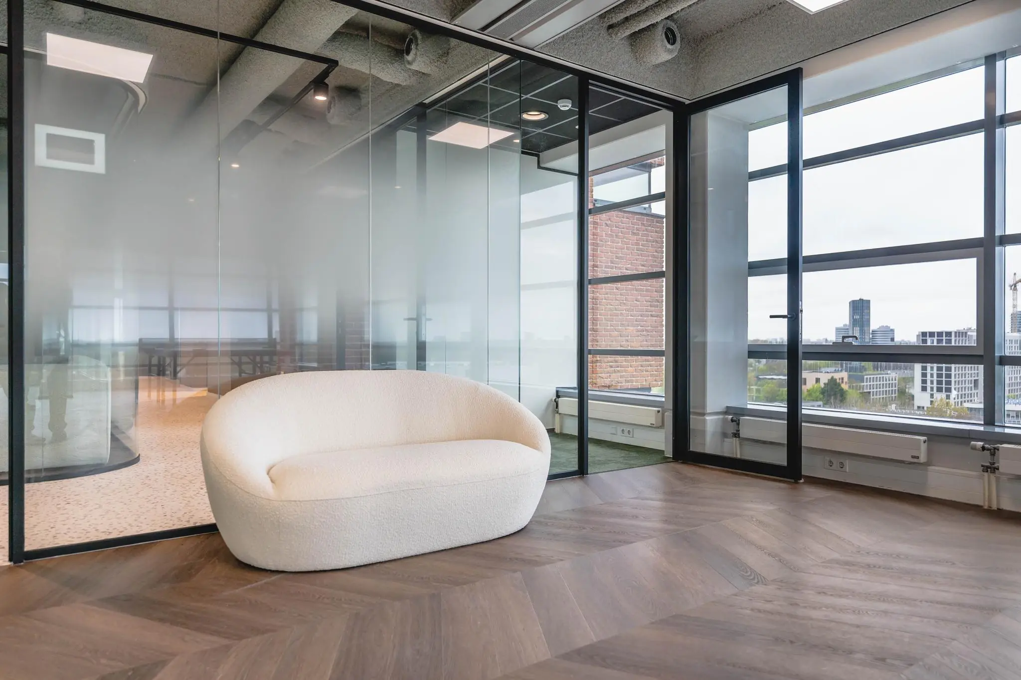 Moderne wachtruimte met een beige designbank, glazen wanden en uitzicht op de skyline vanaf de Joop Geesinkweg.
